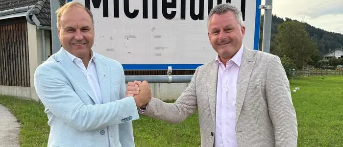 Zwei Männer stehen vor der Ortstafel in Micheldorf, geben sich die Hand und lächeln in die Kamera.