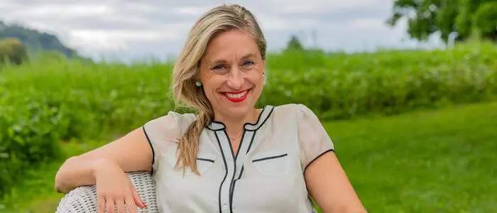 Eine blonde Frau in hellem Kleid sitzt auf einem weißen Stuhl und lacht in die Kamera