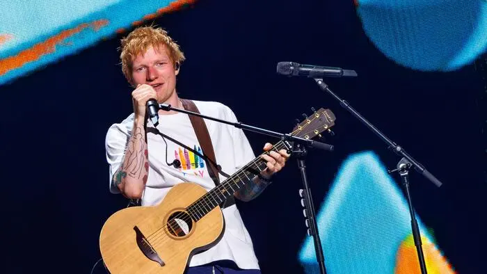 Ed Sheeran hält ein Mikro in der rechten Hand und hat eine Gitarre umgehängt.