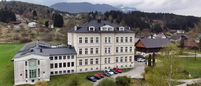 Außenansicht eines mehrstöckigen Schulgebäudes, dahinter grüne Wiesen und hügelige Wälder