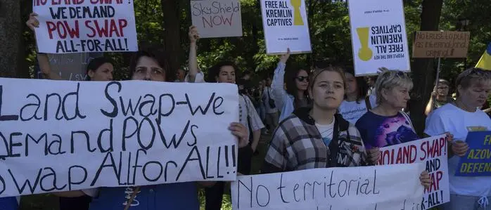 Das Bild zeigt eine Gruppe von Menschen, die im Freien – offenbar in einem Park – an einer Demonstration teilnehmen. Sie halten verschiedene selbstgemachte und gedruckte Plakate hoch, auf denen englische Botschaften stehen. Die Forderungen drehen sich vor allem um den Austausch von Kriegsgefangenen („POW swap“) und die Ablehnung eines „Land Swaps“ (Gebietstausch). | Ukrainerinnen und Ukrainer demonstrieren vor der US-Botschaft in Kiew im Vorfeld des Trump-Putin-Gipfels gegen Gebietsabtretungen