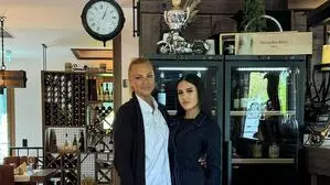 Zwei Frauen stehen in einem Lokal, im Hintergrund sieht man eine Uhr und Weinregale