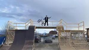 BMX-Sportler Kevin Böck steht mit seinem BMX-Rad in der Hand auf einer hohen Rampe aus Holz, die er selbst gebaut hat