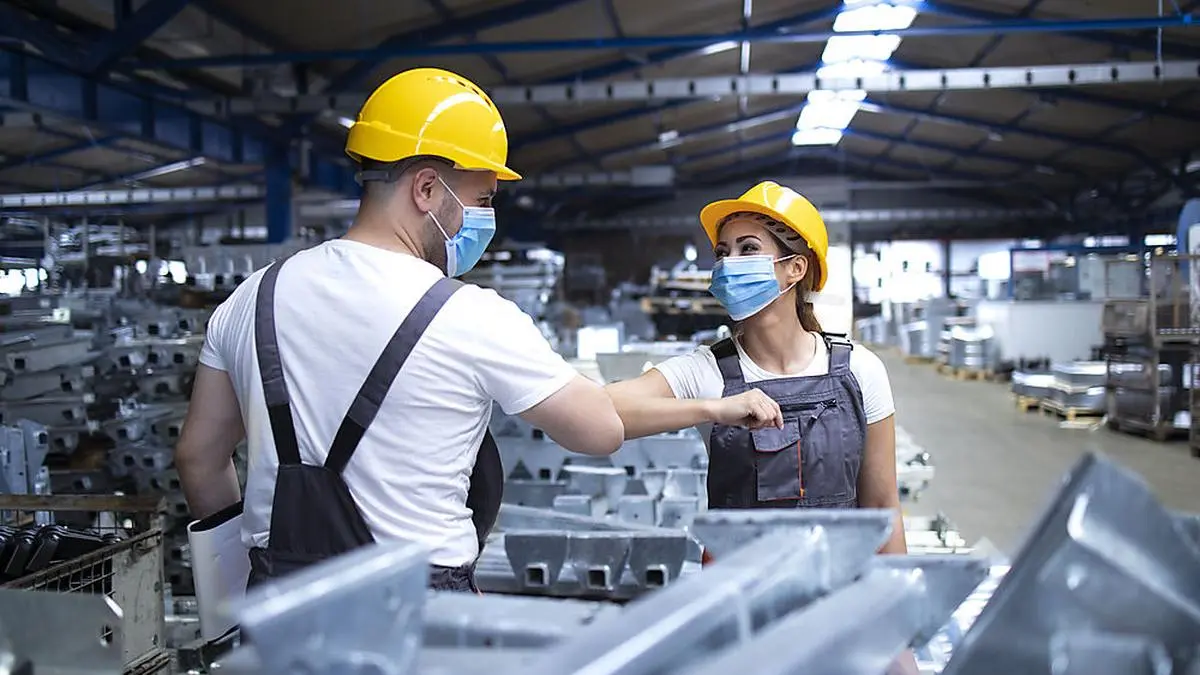 Workers greeting each other with elbow bump due to global corona virus pandemic and danger of infection. Employees standing in factory wearing protection mask.