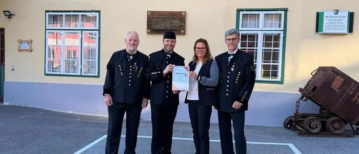 Drei Männer in typischer Bergmannskleidung und eine Frau stehen vor einem gelben Haus auf dem „Bergbau-Museum Bad Bleiberg“ steht