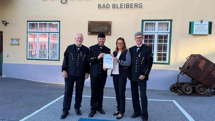 Drei Männer in typischer Bergmannskleidung und eine Frau stehen vor einem gelben Haus auf dem „Bergbau-Museum Bad Bleiberg“ steht