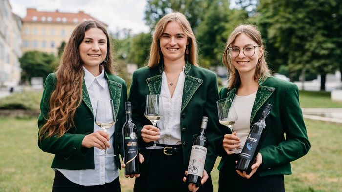 Drei junge Damen in grünen Sakkos lächeln in die Kamera. In ihren Händen halten sie ein Glas Wein und eine Flasche Wein.