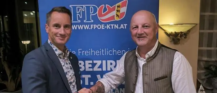 Zwei Männer stehen vor einem FPÖ-Banner, geben sich die Hand und lächeln in die Kamera.