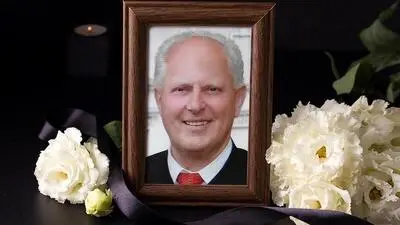 Porträt eines Mannes mit Anzug und grauen Haaren in einem Bilderrahmen, davor weiße Blumen und eine schwarze Schleife