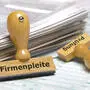 An einem Papierstapel lehnt ein Holzstempel mit dem Aufdruck: Firmenpleite und Prüfung  |  