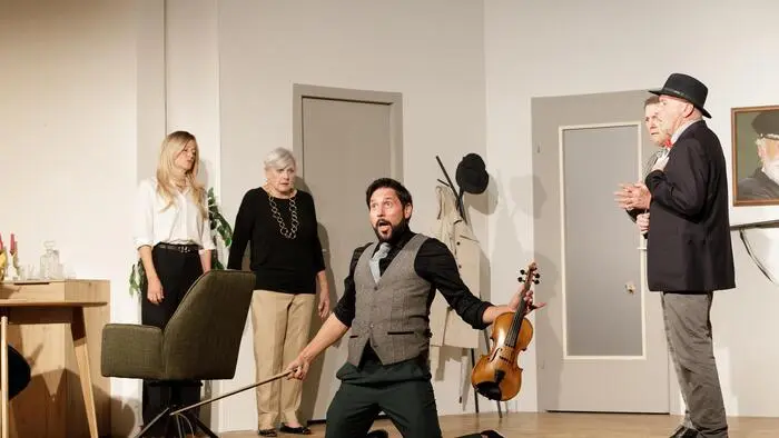 Eine Szene auf einer Theaterbühne: Ein Mann kniet am Boden mit einer Geige in der Hand, daneben stehen Menschen und schauen erschrocken.