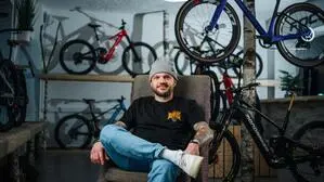 Ein Mann in Jeans, schwarzem Shirt und Haube, sitzt in einem Fahrradgeschäft, im Hintergrund stehen und hängen unterschiedliche Fahrräder