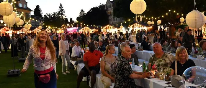 Viele Menschen auf einer Wiese mit geschmückten Bierbänken und -tischen. Es hängen leuchtende Lampions in der Luft