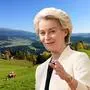 Das Bild zeigt Ursula von der Leyen in eleganter, heller Kleidung, die eine Geste mit der Hand macht. Der Hintergrund ist eine malerische Landschaft mit grünen Wiesen, Hügeln und Bergen im Sonnenlicht. Auf den Wiesen sind Kühe zu sehen, die grasen. | Blick vom Naturpark Zirbitzkogel nahe Neumarkt in die Ferne: Ursula von der Leyen dürfte diese Aussicht kennen