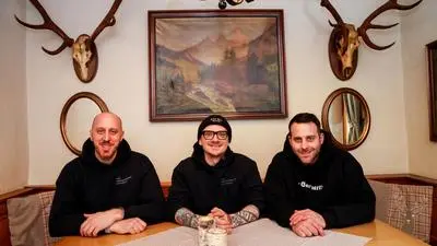 Das Bild zeigt Markus Gallé, Tobias Graf und Lukas Turek, die an einem Holztisch in einer gemütlichen, rustikalen Stube sitzen. Sie tragen schwarze Kapuzenpullover mit Aufdrucken und wirken entspannt und freundlich. Der Mann in der Mitte trägt eine Brille, eine Mütze mit einem aufgedruckten Zielsymbol und hat tätowierte Arme. Die beiden anderen Männer haben kurze Haare, einer von ihnen ist kahl.

Der Raum ist im traditionellen Stil dekoriert, mit einem Landschaftsbild in einem Holzrahmen an der Wand, zwei Spiegeln auf beiden Seiten und zwei Hirschgeweihen mit Schädeln darüber. Die Sitzbank hat Kissen mit Hirschmotiven, was die rustikale Atmosphäre unterstreicht. Auf dem Tisch liegt eine gestreifte Tischdecke, und es stehen zwei kleine, aus Baumstämmen gefertigte Kerzenhalter darauf.

Die Beleuchtung ist warm, und die Gesamtstimmung des Bildes wirkt freundlich und einladend.