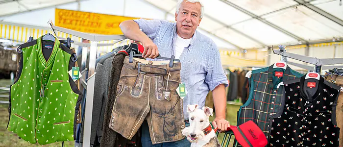 Mann mit weißen Haaren steht vor Trachtenmode, neben ihm ein Hund