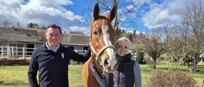Auf dem Foto sieht man einen dunkelhaarigen Mann und eine blonde Frau, die ein braunes Pferd mit weißer Blässe zwischen sich am Zügel hält. Im Hintergrund sieht man eine grüne Wiese und ein Stallgebäude mit weißem Zaun. | Heimo und Bettina Kendlbacher mit ihrem Pferd Don Alfredo