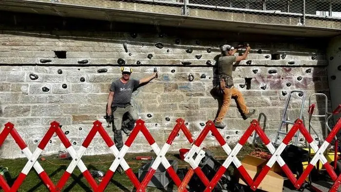 Zwei Männer montieren Klettergriffe an einer Wand, davor steht eine Absperrung
