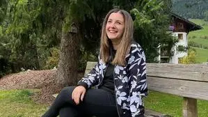 Eine Frau mit dunklen Haaren trägt eine schwarze Hose und eine Jacke mit Blumen, dabei sitzt sie auf einer Holzbank und lacht