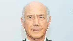 Ein Mann mit weißen Haaren und braunem Anzug lacht in die Kamera