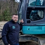 Ein Mann mit kurzen schwarzen Haaren steht vor einem blauen Bagger | Kevin Michael Wurm plant sein Unternehmen weiter auszubauen 