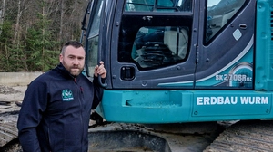 Ein Mann mit kurzen schwarzen Haaren steht vor einem blauen Bagger | Kevin Michael Wurm plant sein Unternehmen weiter auszubauen 