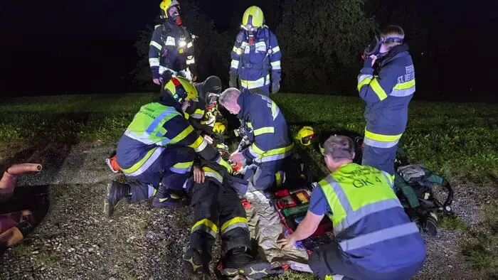 Man sieht ein halbes Dutzend Feuerwehrmänner, einige davon mit Atemschutz, die eine Person auf den Boden legen.