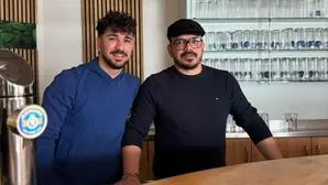 Zwei dunkelhaarige Männer stehen hinter einer Theke und lachen in die Kamera. Der linke trägt einen blauen Pullover, der rechte einen schwarzen Pullover und eine Kappe | Die Brüder Simone und Carmelo Lombardo (von links)