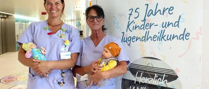 Zwei Pflegerinnen mit Puppen im Arm stehen vor einem Schild mit der Aufschrift „75 Jahre Kinder- und Jugendheilkunde“