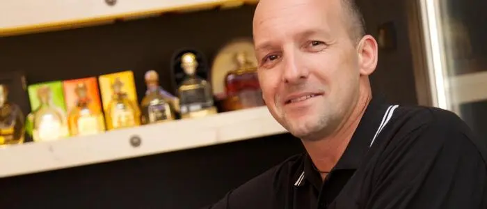 Mann mit Glatze und schwarzem Poloshirt sitzt lässig vor einem Regal | Johannes Schuschu, 58, baut aktuell die Hotelkette „Ensana“ in Österreich und Deutschland auf