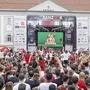 Viele Menschen starren auf eine große Leinwand, auf der ein Fußballspiel übertragen wird.