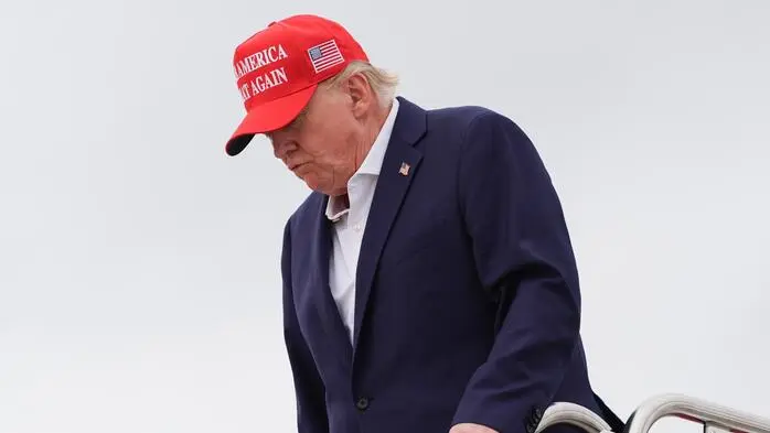 Donald Trump hält sich an einem Handlauf fest. Er trägt ein Hemd, ein Sakko und eine rote Kappe.