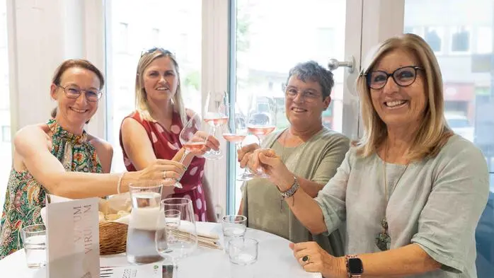 Vier Frauen stoßen mit einem Glas Wein an und lächeln in die Kamera.