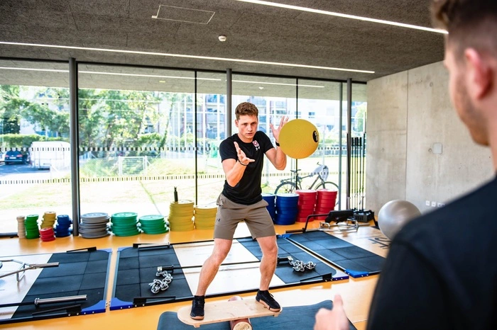 Beinachsen Stabilitätsübung auf dem IndoBoard mit Medizinballwürfen: Training des Gleichgewichtssinns mit erhöhter Anforderung an den Rumpf durch die seitliche Fang- und Wurfbewegung mit dem Medizinball.