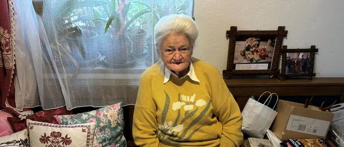 Sieglinde Talker sitzt auf einer Eckbank in ihrem Haus, auf dem Tisch ist ihre Handwerkskunst ausgebreitet. Sie selbst hat graue hochgesteckte Haare und trägt einen gelben Pullover
