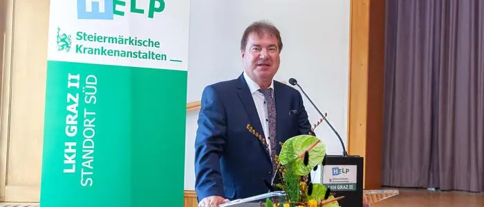Ein Mann im dunkelblauen Anzug steht vor einem Stehpult und spricht gerade in ein Mikrofon.