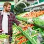 Handsome man with shopping trolley touching tomato in order to define its quality in fruit and vegetable department of supermarket