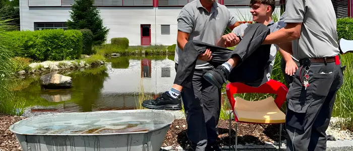 Eine Wanne steht vor einem Teich, zwei Männer tragen den Lehrling