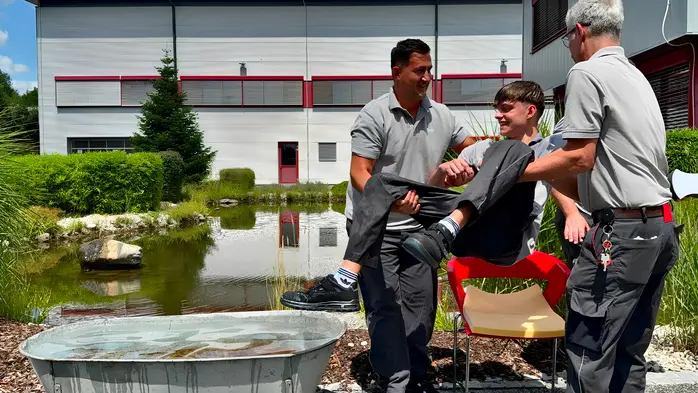 Eine Wanne steht vor einem Teich, zwei Männer tragen den Lehrling