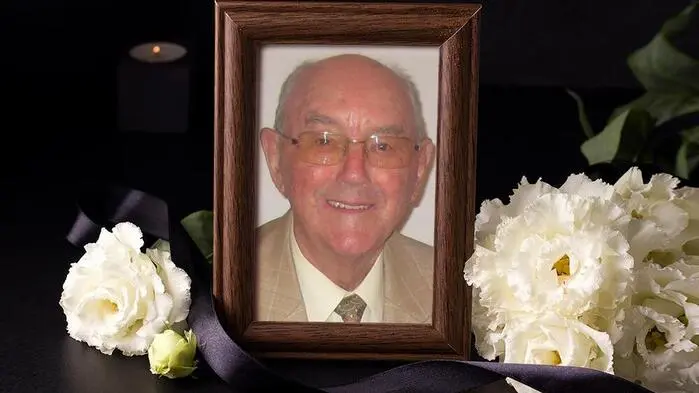 Ein Bild von einem Mann mit  Brille in einem dunklen Bilderrahmen. Darum sind weiße Blumen drapiert