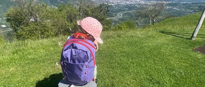 Ein Kleinkind mit Rucksack und Kappe auf einem Schaukelpferd, im Hintergrund Blick auf Bergpanorama und eine Stadt