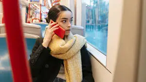 Das Foto zeigt eine Frau, die in einem Öffentlichen Verkehrsmittel sitzt und sich eine Handy ans Ohr hält.