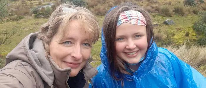 Eine blonde Frau und eine dunkelhaarige Jugendliche lachen in die Kamera, im Hintergrund sind ein See und Berge zu sehen