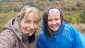 Eine blonde Frau und eine dunkelhaarige Jugendliche lachen in die Kamera, im Hintergrund sind ein See und Berge zu sehen