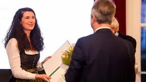 Frau mit dunklen, langen Locken im Dirndl bekommt von zwei Männern eine Urkunde  überreicht