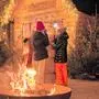 Mehrere Personen an einer Feuerschale, dahinter eine Holzhütte und Christbäume | Der Orasch‘sche Adventmarkt in Tainach endet – die Christbäume suchen Abnehmer