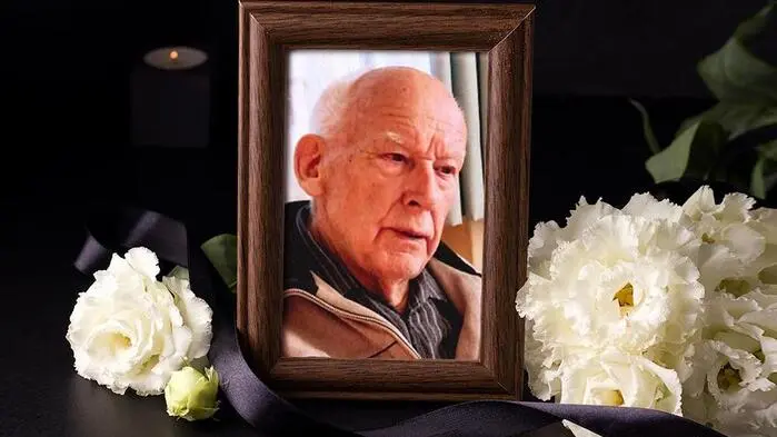 Ein Bild von einem älteren Herren umrahmt von weißen Blumen und schwarzen Schleifen
