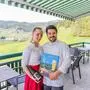 Eine Frau und ein Mann lächeln auf einer Terrasse in die Kamera.