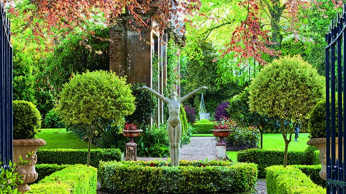 GRANGE COURT GUERNSEY: VIEW THROUGH METAL GATES TO SCULPTURE ´WELCOME THE NEW YEAR´ BY EVERARD MEYNELL IN BOX BEDS WITH LAWN AND COPPER BEECH BEHIND