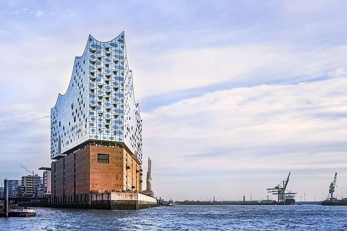 Und das beeindruckende, von drei Seiten vom Wasser der Elbe umgebene Gebäude wurde zum Touristenmagneten und zum neuen, weithin sichtbaren Wahrzeichen der Stadt. Auf einem Backsteinsockel, einem ehemaligen Kaispeicher, ragt der gläserne Neubau mit seiner geschwungenen Dachlandschaft bis zu 110 Meter in den Himmel, erbaut an der westlichen Spitze der HafenCity und in Nachbarschaft zu der mit Backsteinen erbauten und mittlerweile zum Unesco-Welterbe zählenden Speicherstadt.